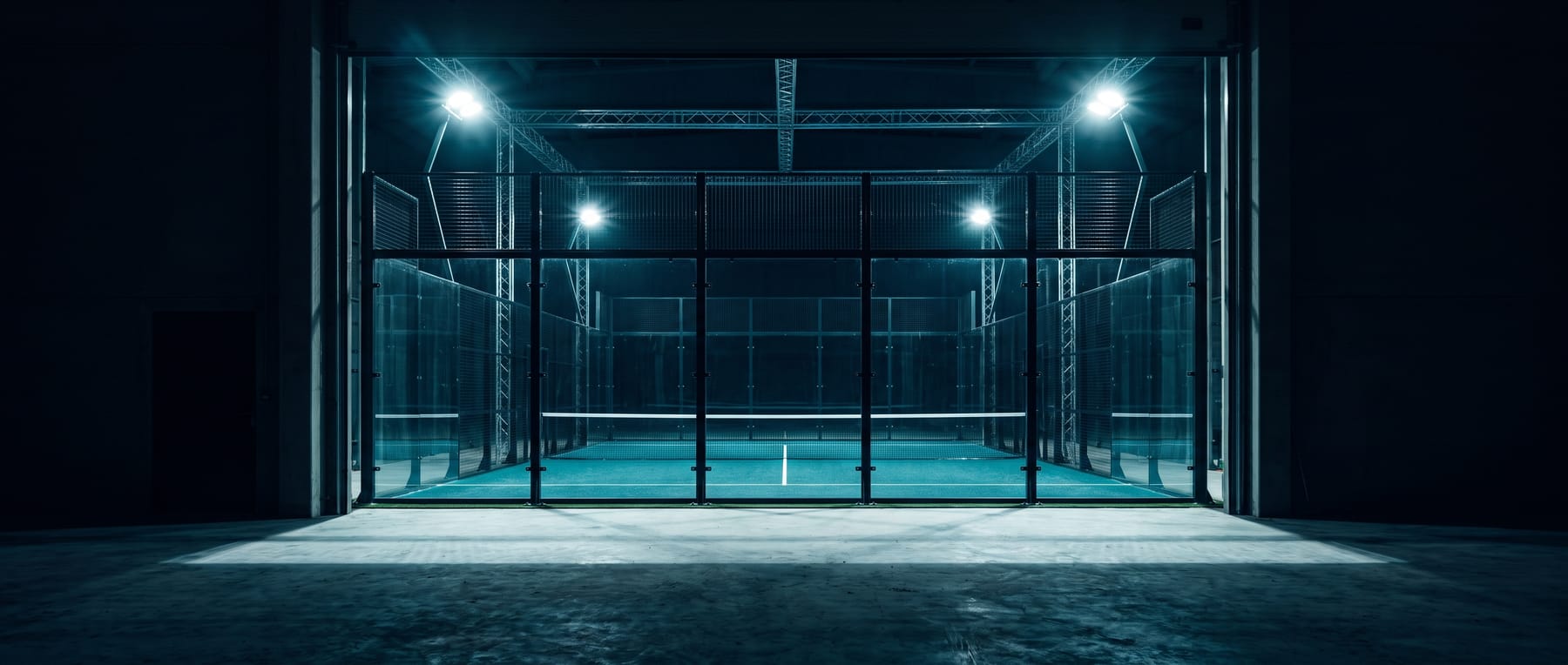 The same padel court at night, blazing under eight floodlights so the glass enclosure glows like a lantern in an otherwise black warehouse.