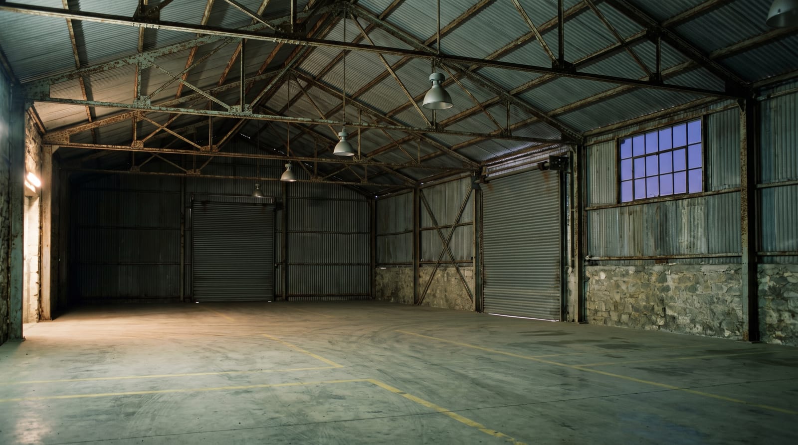 The interior of the warehouse at dusk: rusted steel roof trusses, corrugated cladding, a closed roller shutter and a bare concrete floor marked with faded yellow lines.